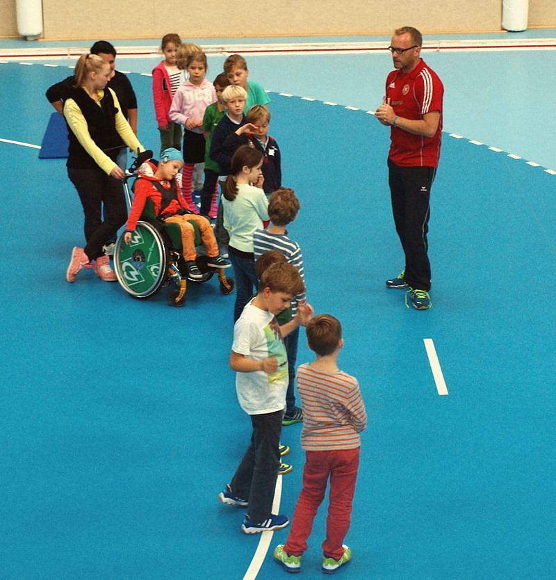 Kinder machen mit einem Trainer Bewegungsspiele in der Sporthalle, ein Kind im Rollstuhl ist dabei